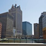 Downtown West Allis, WI skyline with historic and modern buildings along a riverwalk on a clear, sunny day.