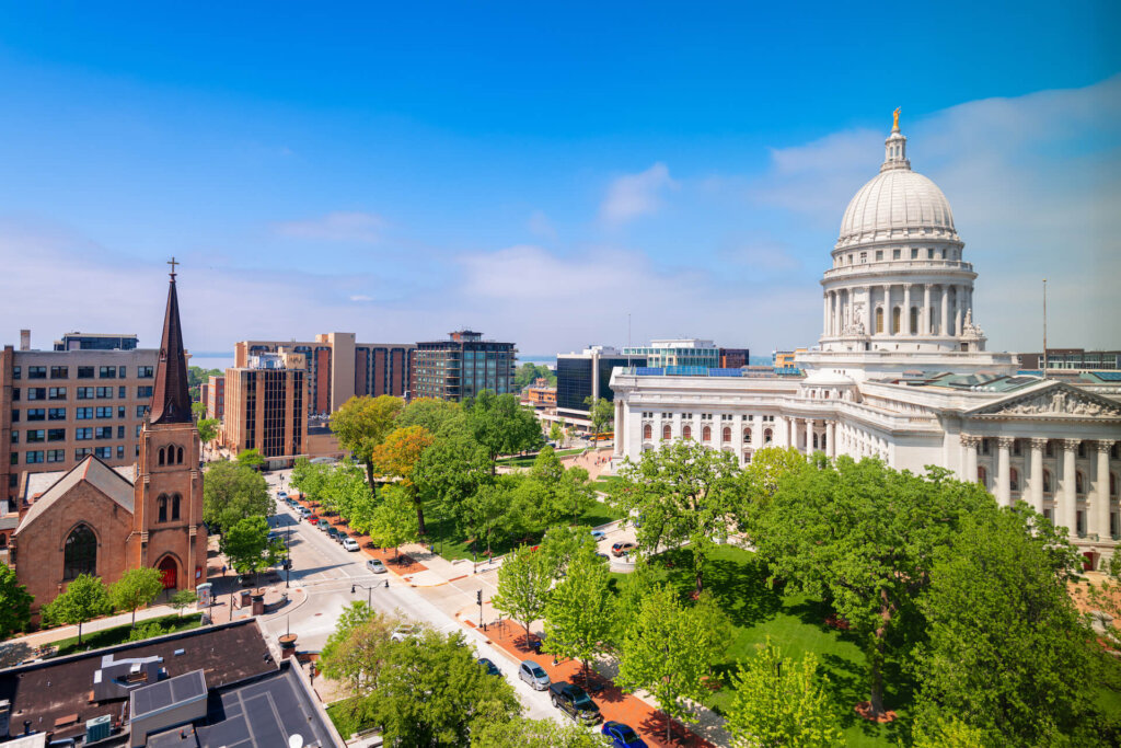 A sunny view of downtown Madison, Wisconsin, featuring the Wisconsin State Capitol building surrounded by green trees and nearby city buildings.