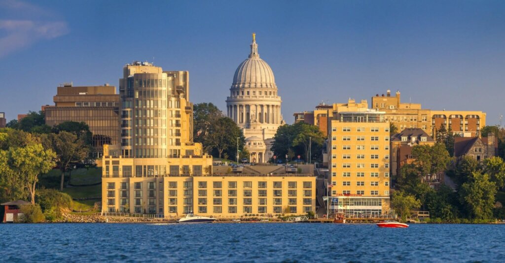 Skyline view of downtown Madison, Wisconsin with the State Capitol building centered behind modern waterfront buildings along Lake Mendota.