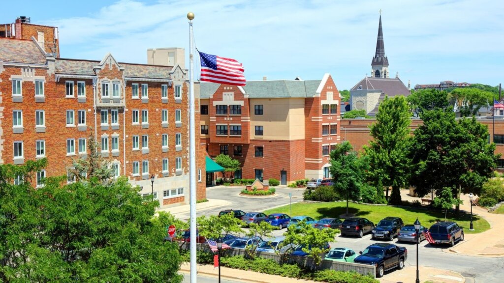 Downtown Waukesha, Wisconsin featuring historic brick buildings, a U.S. flag, parked cars, and a church steeple in the background on a sunny day.