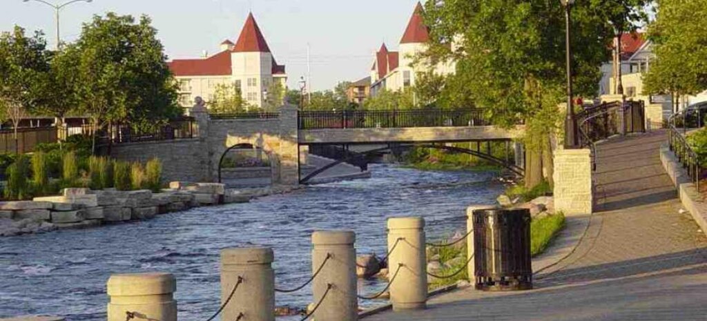 Scenic riverwalk in downtown Waukesha with stone bridge, flowing river, and historic buildings in the background.