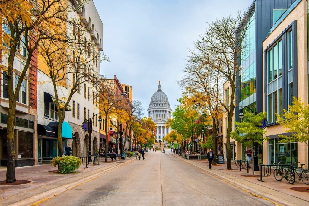 Downtown Madison, Wisconsin in autumn with the State Capitol building in view, framed by colorful trees and modern storefronts.