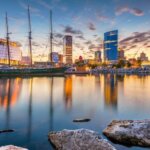 Downtown Milwaukee skyline at sunset with city lights reflecting on Lake Michigan, featuring the Discovery World building and sailboat.
