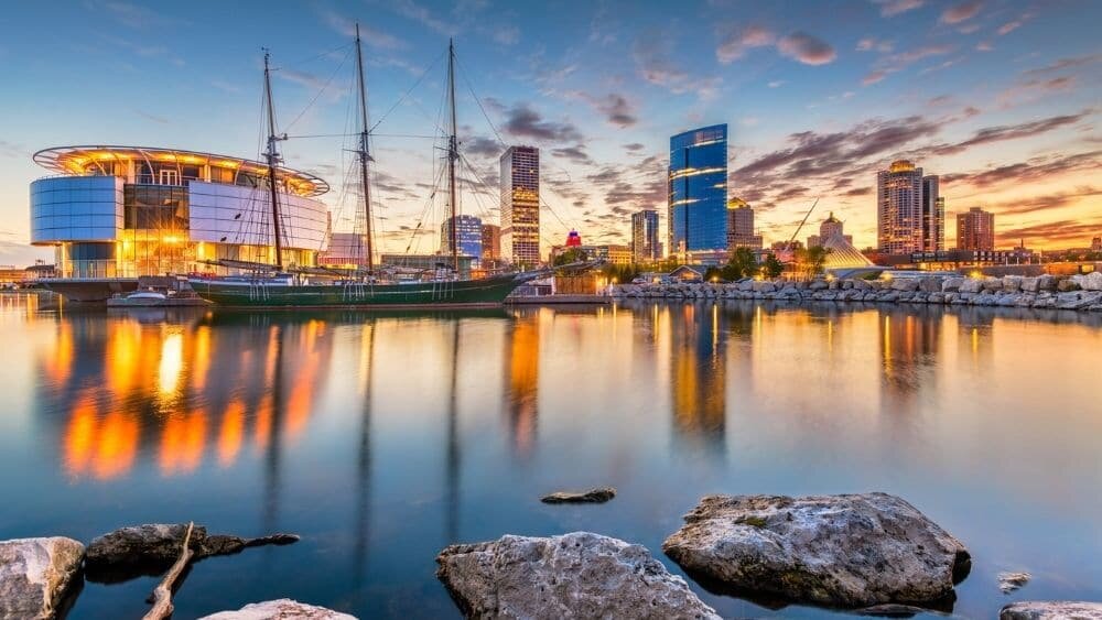 Downtown Milwaukee skyline at sunset with city lights reflecting on Lake Michigan, featuring the Discovery World building and sailboat.