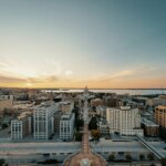 Aerial view of downtown Madison, Wisconsin at sunset, featuring the Wisconsin State Capitol and surrounding city buildings with lakes in the background.