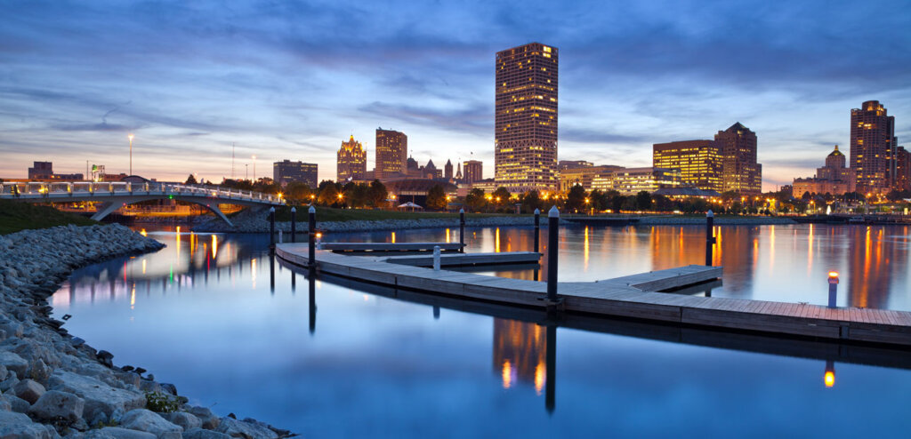 Milwaukee skyline at dusk reflected in calm waters with a dock in the foreground.