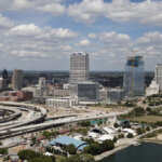 Aerial view of downtown Milwaukee with high-rise buildings, highways, and Lake Michigan on a partly cloudy day.