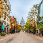State Street in downtown Madison, Wisconsin, lined with shops and trees in fall, leading to the Wisconsin State Capitol building in the distance.