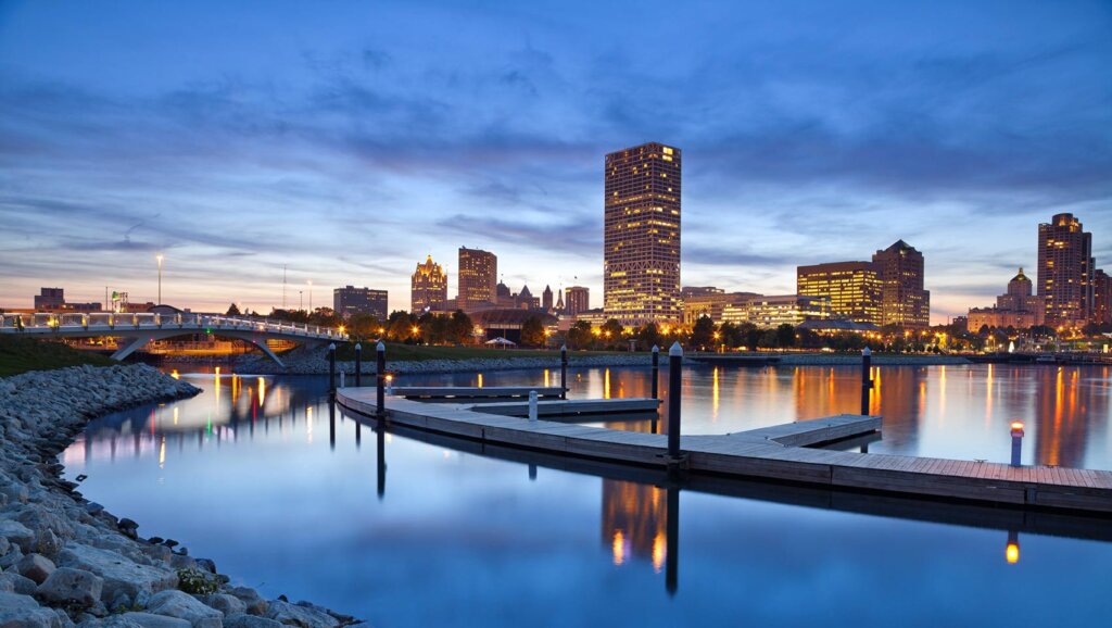 Milwaukee city skyline at dusk with reflections on the river and a modern dock in the foreground.