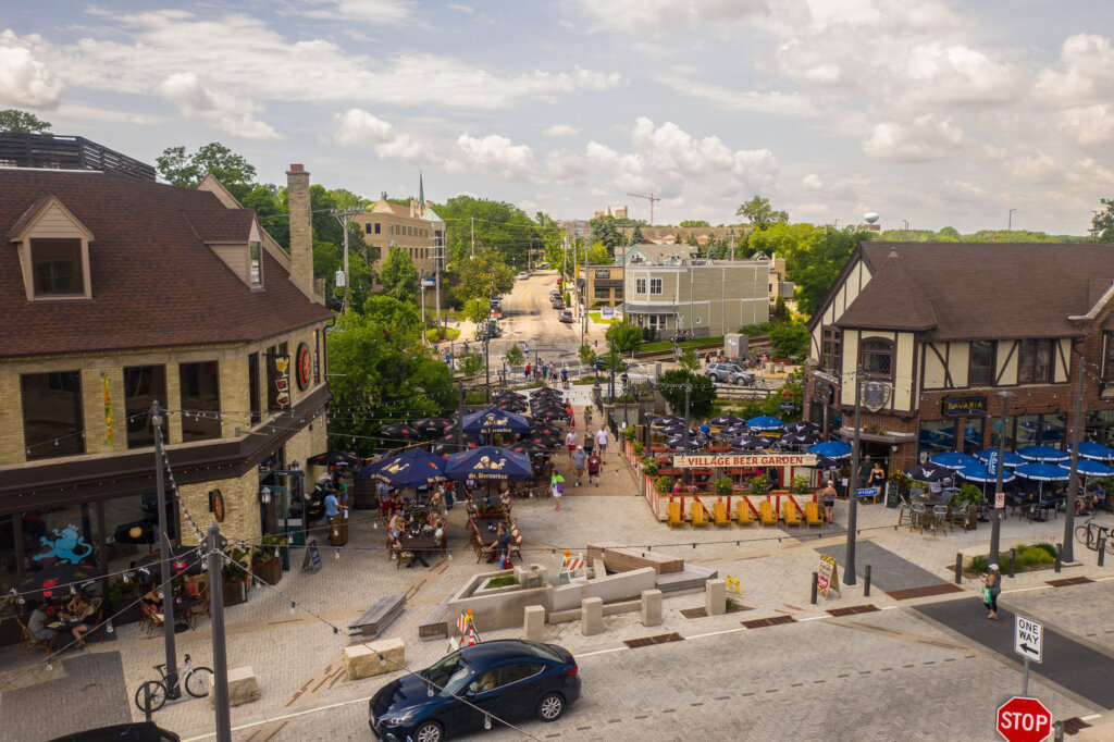 A lively outdoor beer garden in Wauwatosa's Village district, surrounded by people dining under umbrellas, historic buildings, and a charming pedestrian street.