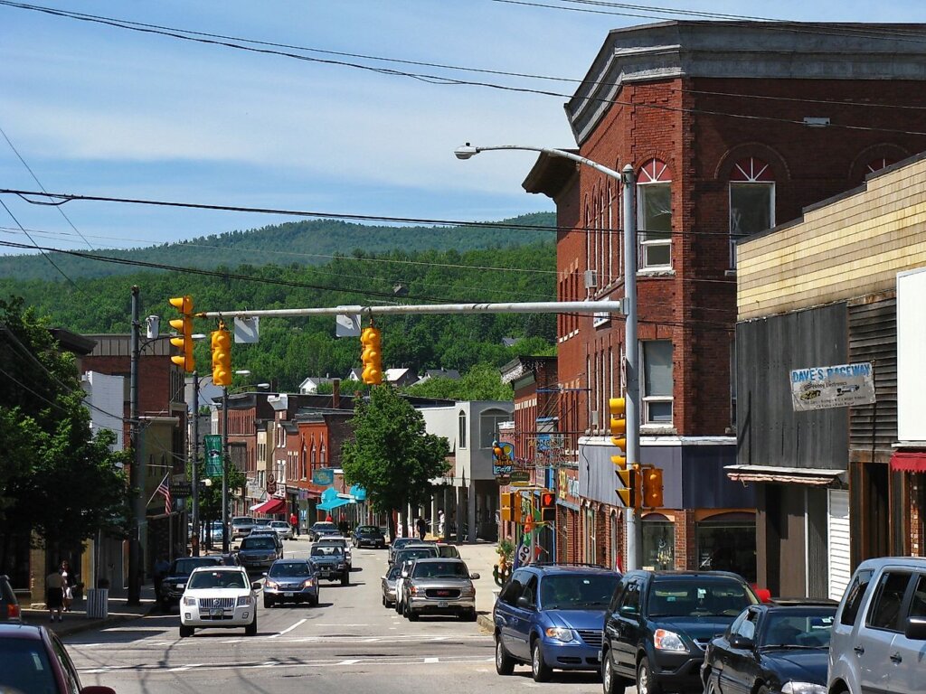 Busy small-town street in New Berlin with cars, historic brick buildings, shops, and green hills in the background.
