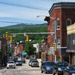 Busy small-town street in New Berlin with cars, historic brick buildings, shops, and green hills in the background.