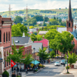 A vibrant downtown street in West Allis with historic brick buildings, a church steeple, shops, and green hills in the background.