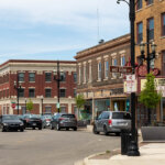 Downtown Kenosha street view with historic brick buildings, shops, cars parked along the road, and classic black street lamps on a sunny day.