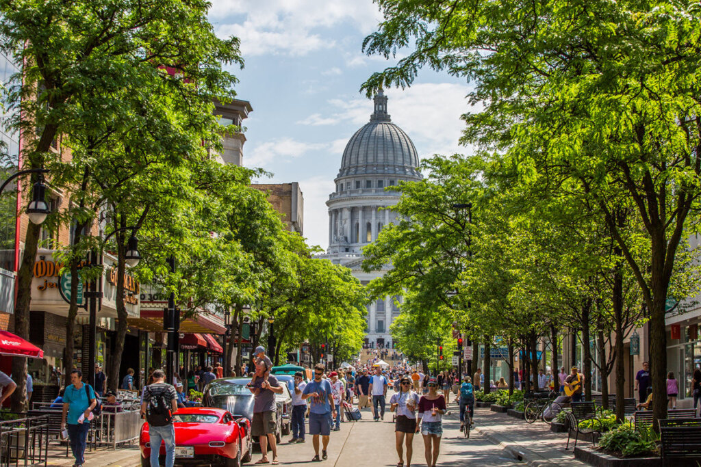 Bustling downtown Madison, Wisconsin street lined with trees, cafes, and pedestrians, with the Wisconsin State Capitol building visible in the background on a sunny day.