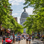 Bustling downtown Madison, Wisconsin street lined with trees, cafes, and pedestrians, with the Wisconsin State Capitol building visible in the background on a sunny day.