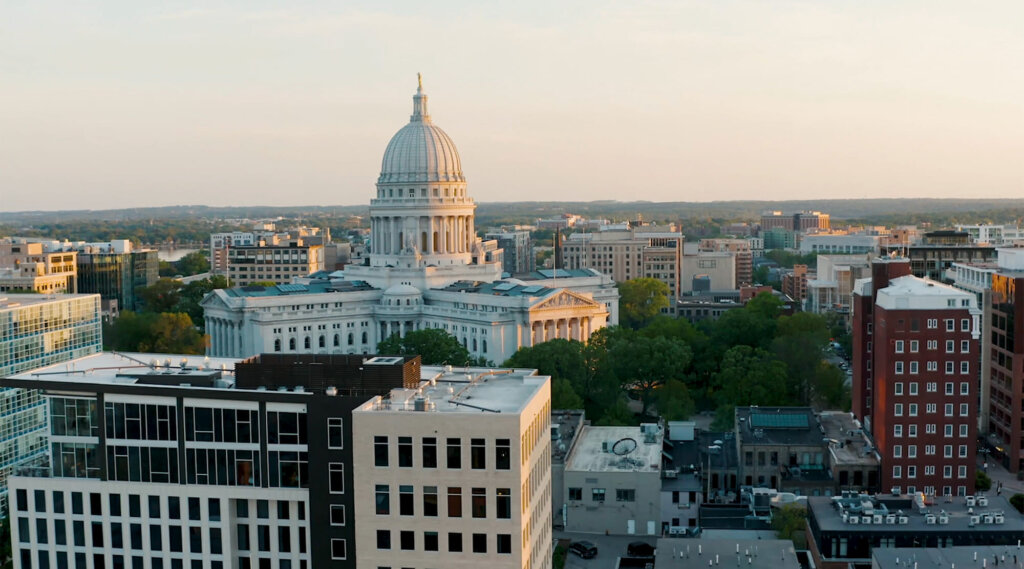 Aerial view of downtown Madison, Wisconsin, featuring the Wisconsin State Capitol building surrounded by modern offices and residential buildings at sunset.