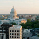 Aerial view of downtown Madison, Wisconsin, featuring the Wisconsin State Capitol building surrounded by modern offices and residential buildings at sunset.