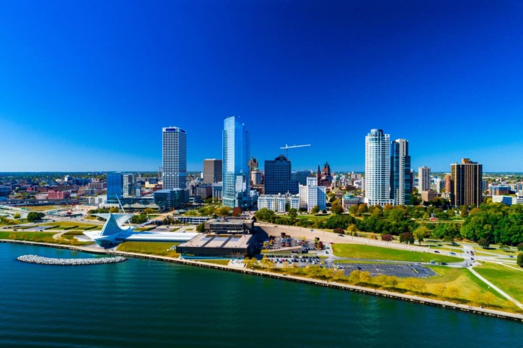 Aerial view of downtown Milwaukee, Wisconsin, showing modern skyscrapers, the Milwaukee Art Museum, and Lake Michigan under a bright blue sky.
