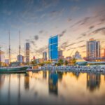 Downtown Milwaukee skyline at sunset with sailboats docked along the waterfront and modern buildings reflecting on the calm water.