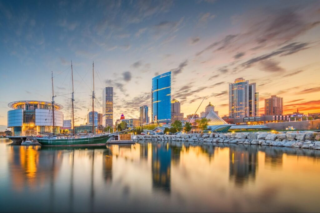 Downtown Milwaukee skyline at sunset with sailboats docked along the waterfront and modern buildings reflecting on the calm water.
