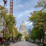 View of downtown Madison’s State Street lined with trees and shops, leading toward the Wisconsin State Capitol under a partly cloudy sky.