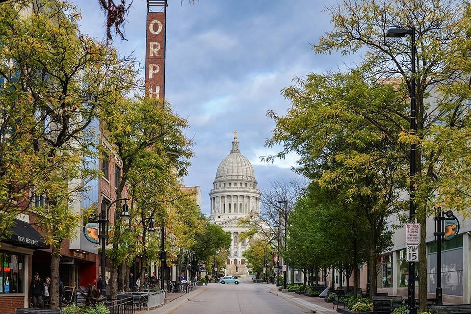 View of downtown Madison’s State Street lined with trees and shops, leading toward the Wisconsin State Capitol under a partly cloudy sky.