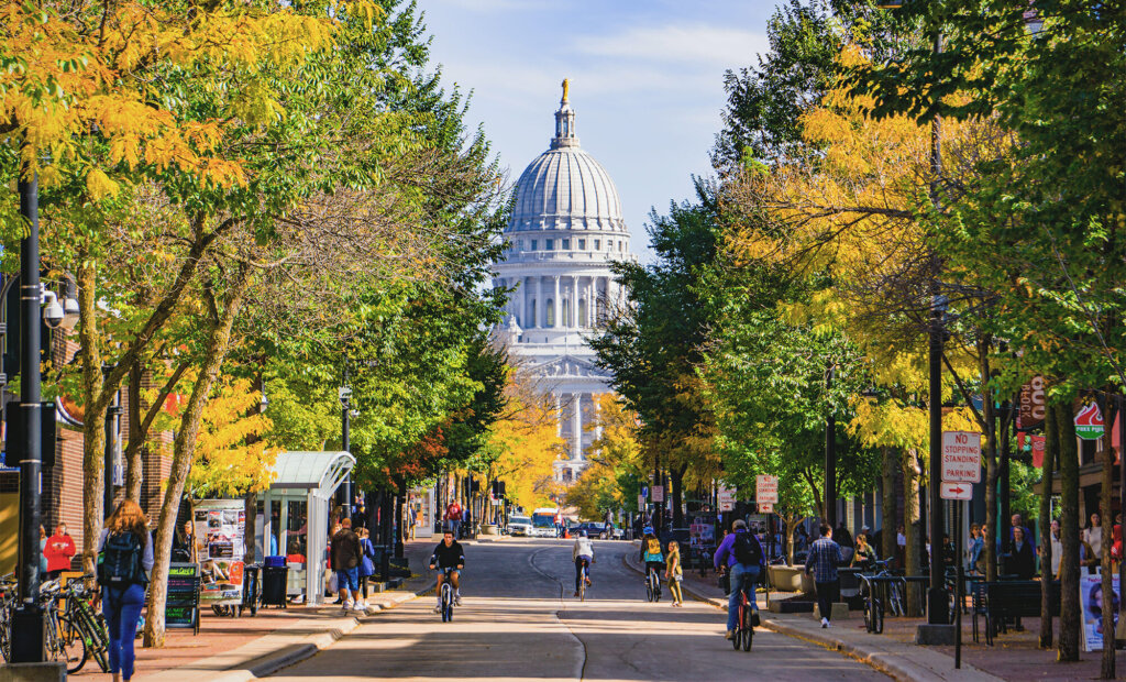 Downtown Madison, Wisconsin showing the State Capitol framed by colorful trees and people biking along State Street on a bright fall day.