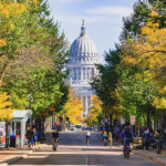 Downtown Madison, Wisconsin showing the State Capitol framed by colorful trees and people biking along State Street on a bright fall day.