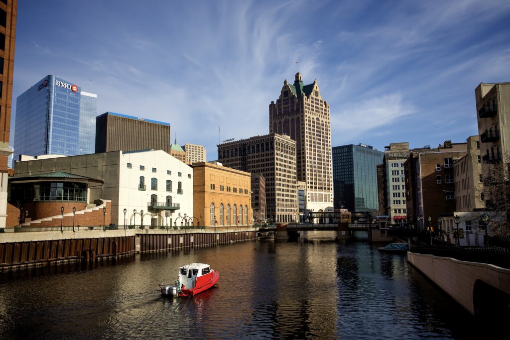 Downtown Milwaukee skyline reflected in the river on a sunny day, featuring modern office buildings and a small red-and-white boat cruising along the calm water.