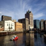 Downtown Milwaukee skyline reflected in the river on a sunny day, featuring modern office buildings and a small red-and-white boat cruising along the calm water.