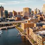 Aerial view of downtown Milwaukee showing the river, bridges, and high-rise buildings under a clear sky at sunset.