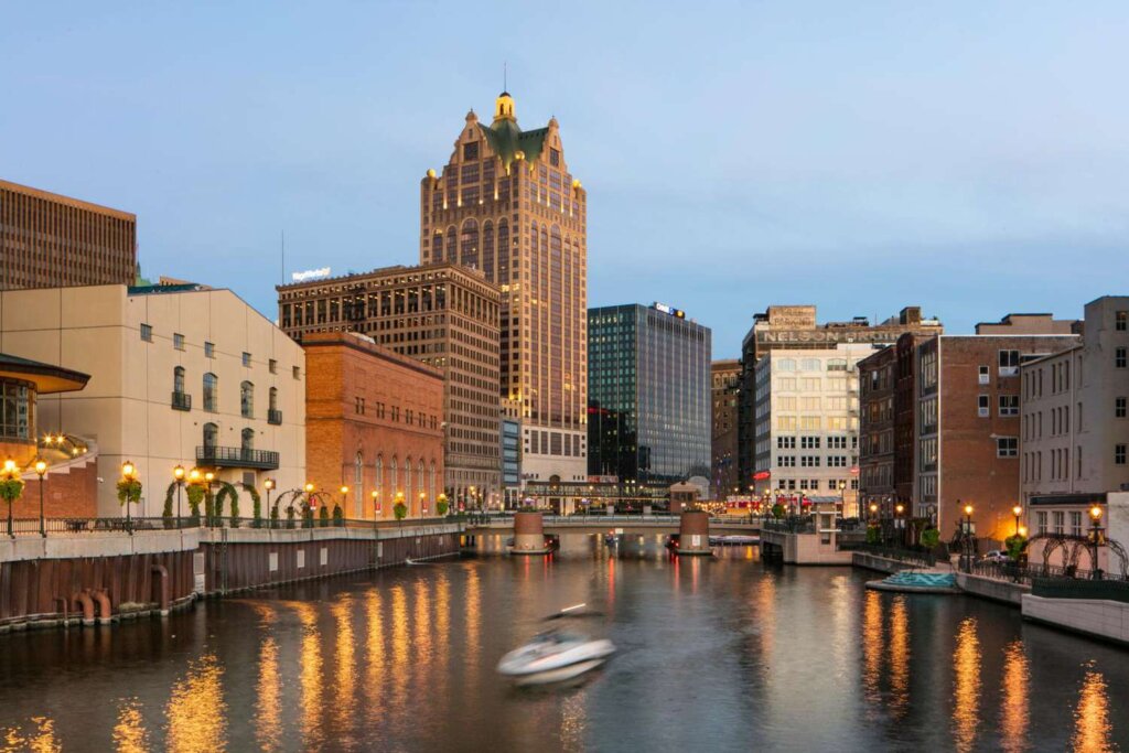 Downtown Milwaukee skyline at dusk with buildings reflecting on the Milwaukee River and a small boat moving across the water.