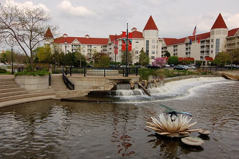 Scenic view of downtown Waukesha featuring a small waterfall, decorative water sculpture, and buildings with red roofs in the background.
