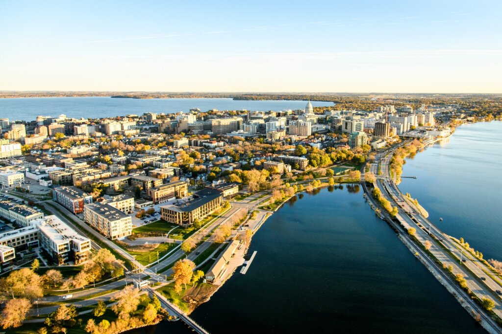 Aerial view of downtown Madison, Wisconsin, showcasing lakeside roads, waterfront parks, and city buildings under clear golden sunlight.
