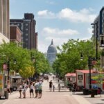 Street view of downtown Madison, Wisconsin, with people walking between shops and food carts toward the Wisconsin State Capitol on a sunny day.