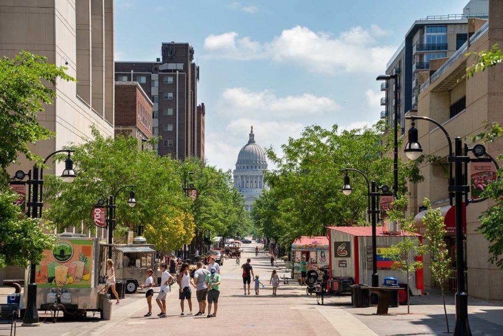 Street view of downtown Madison, Wisconsin, with people walking between shops and food carts toward the Wisconsin State Capitol on a sunny day.