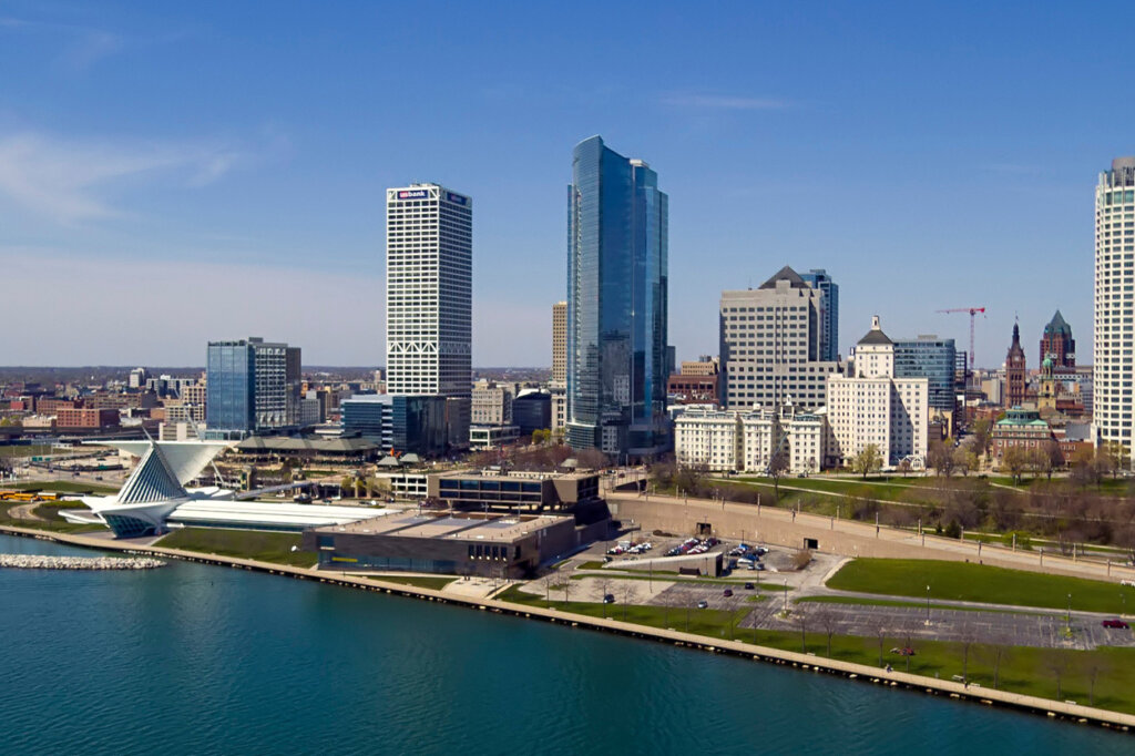 A scenic aerial view of downtown Milwaukee featuring tall modern buildings along the waterfront on a clear, sunny day.