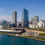 A scenic aerial view of downtown Milwaukee featuring tall modern buildings along the waterfront on a clear, sunny day.