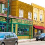 Colorful storefronts and small businesses along a busy street in West Allis, Wisconsin, with cars parked and driving past.