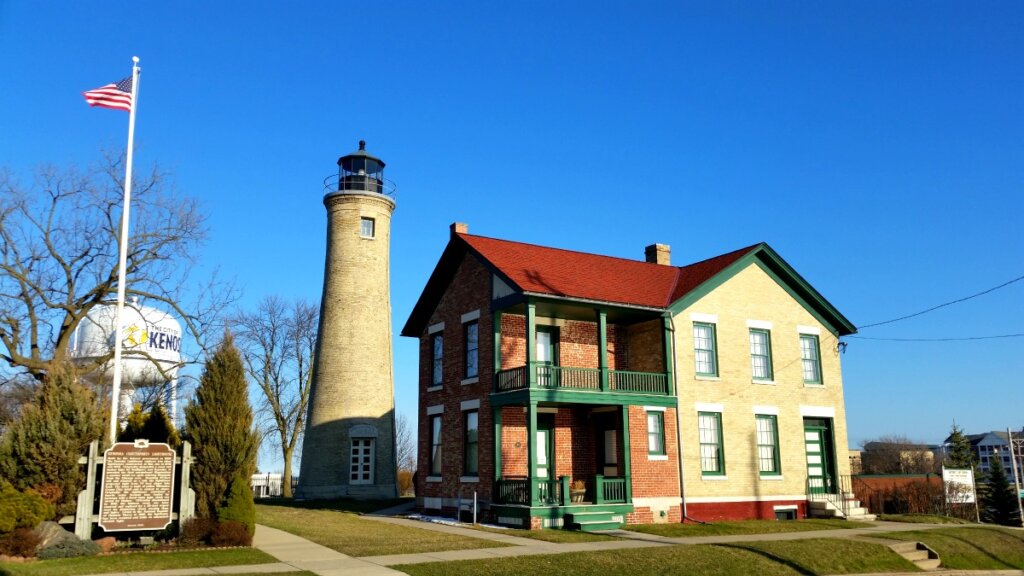 Historic Southport Lighthouse and keeper’s house in Kenosha under a clear blue sky, with an American flag and surrounding greenery.