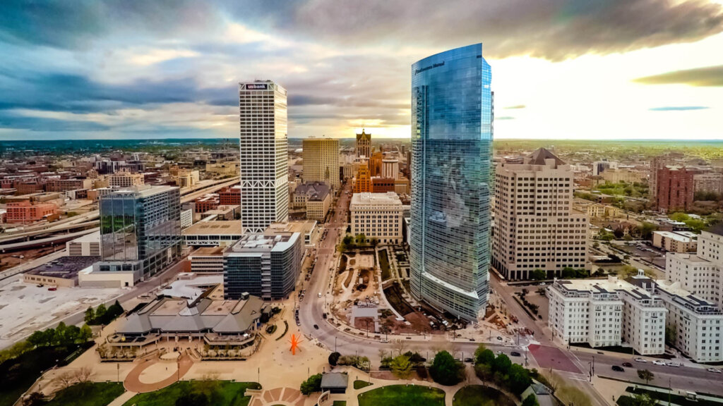 Aerial view of downtown Milwaukee featuring tall modern skyscrapers, city streets, and a dramatic sunset sky.