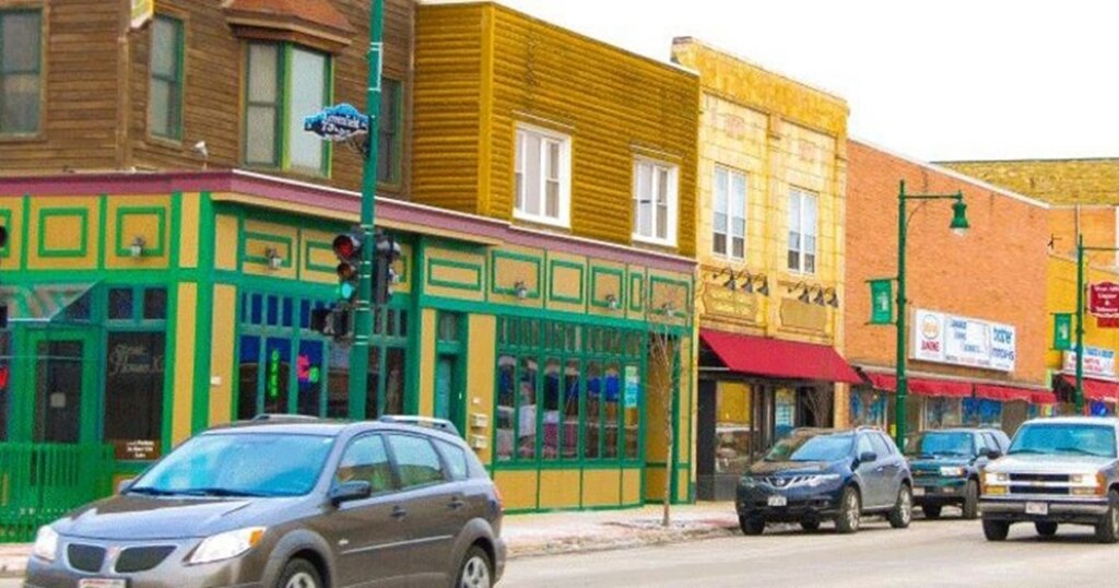 Colorful storefronts along a busy street in West Allis, Wisconsin, with cars parked and driving past local shops.