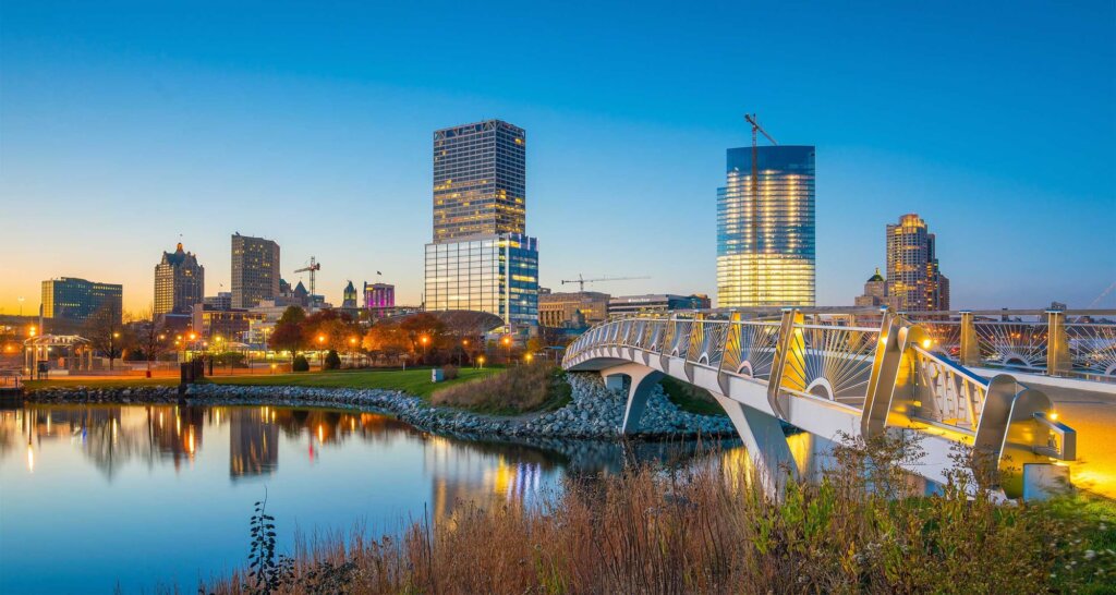 Milwaukee skyline at sunset with modern high-rise buildings reflecting on the calm waterfront and a brightly lit pedestrian bridge in the foreground.