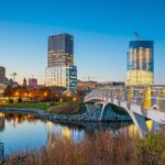 Milwaukee skyline at sunset with modern high-rise buildings reflecting on the calm waterfront and a brightly lit pedestrian bridge in the foreground.