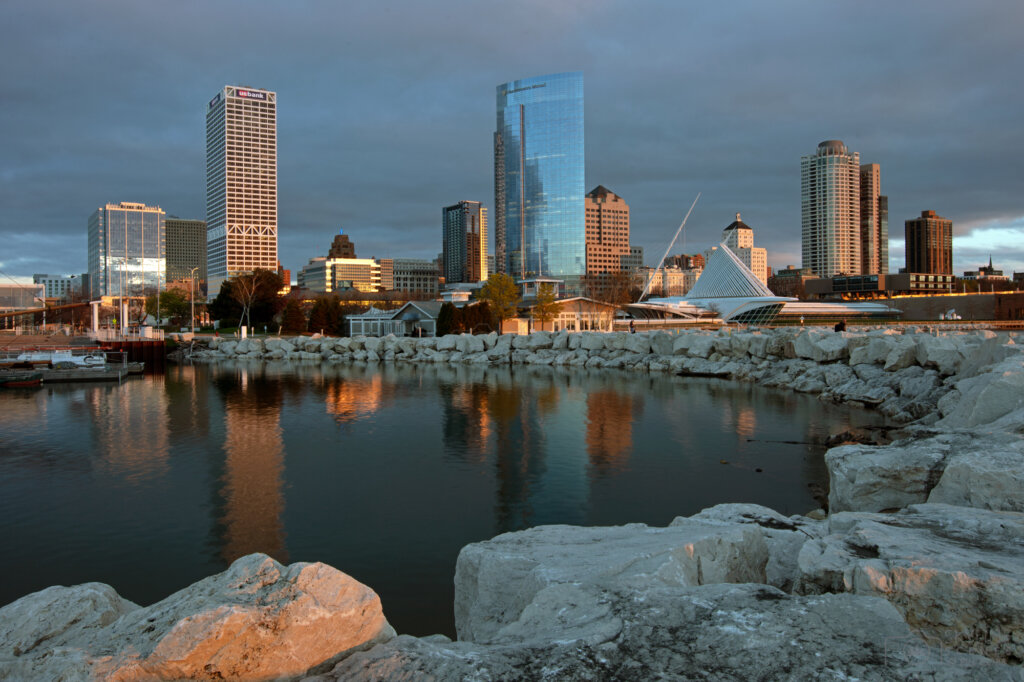 Downtown Milwaukee skyline at sunset reflected on Lake Michigan, featuring modern high-rise buildings and the Milwaukee Art Museum.