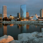 Downtown Milwaukee skyline at sunset reflected on Lake Michigan, featuring modern high-rise buildings and the Milwaukee Art Museum.