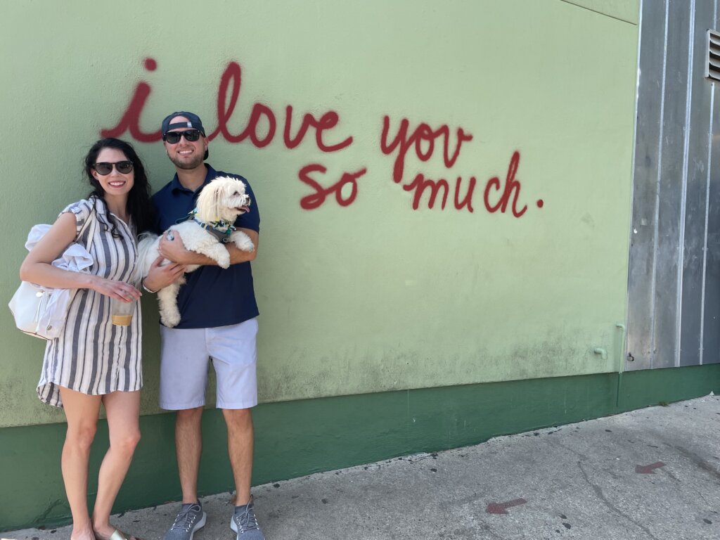 ATX Home Buyer owners, Taylor and Ashley with their dog, Teddy, in front of the famous 'I love you so much' mural in Austin.