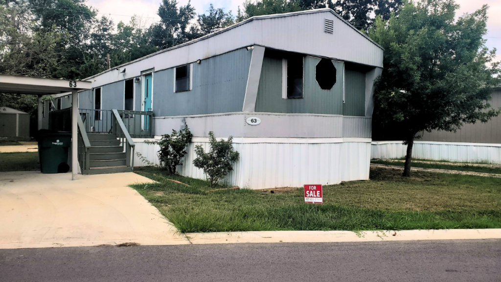 Gray singlewide mobile home trailer in a park, with a For Sale sign in front of it.