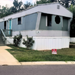 Gray singlewide mobile home trailer in a park, with a For Sale sign in front of it.